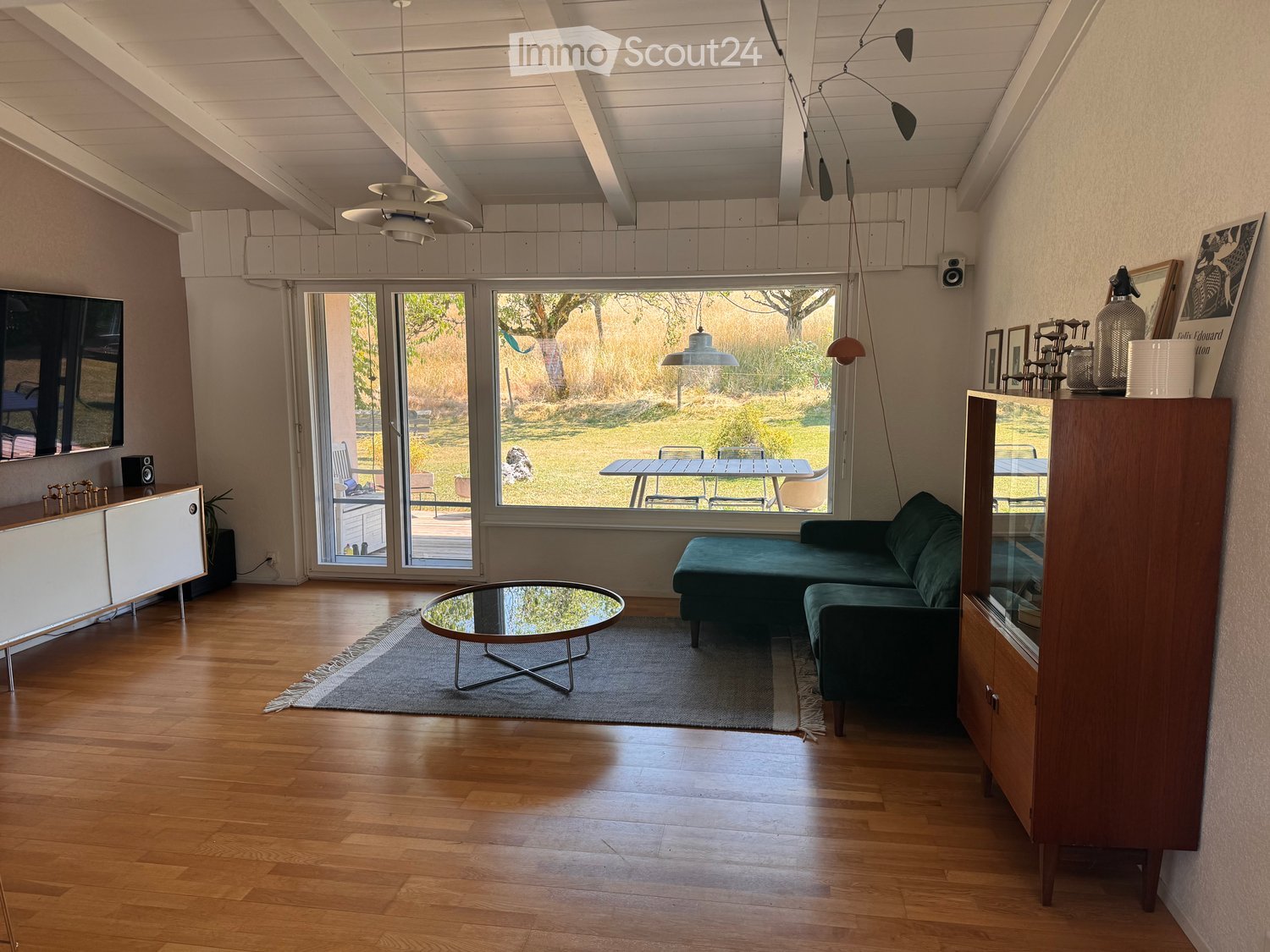 Modern living room with wooden flooring, large windows, a green sofa, a round glass coffee table, and a wooden cabinet with a mirror.