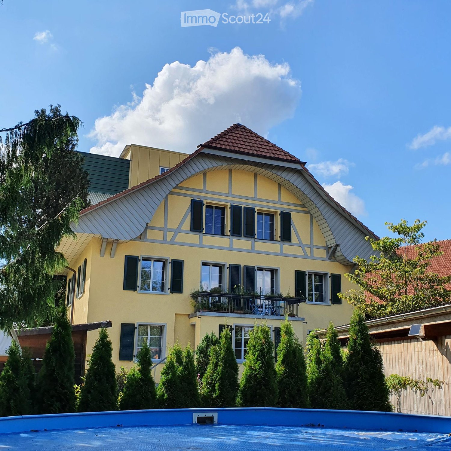 Two-story yellow house with a red-tiled roof, multiple windows with shutters, and a small balcony with outdoor furniture. The house is surrounded by tall, neatly trimmed evergreen trees and a swimming pool is visible in the foreground.