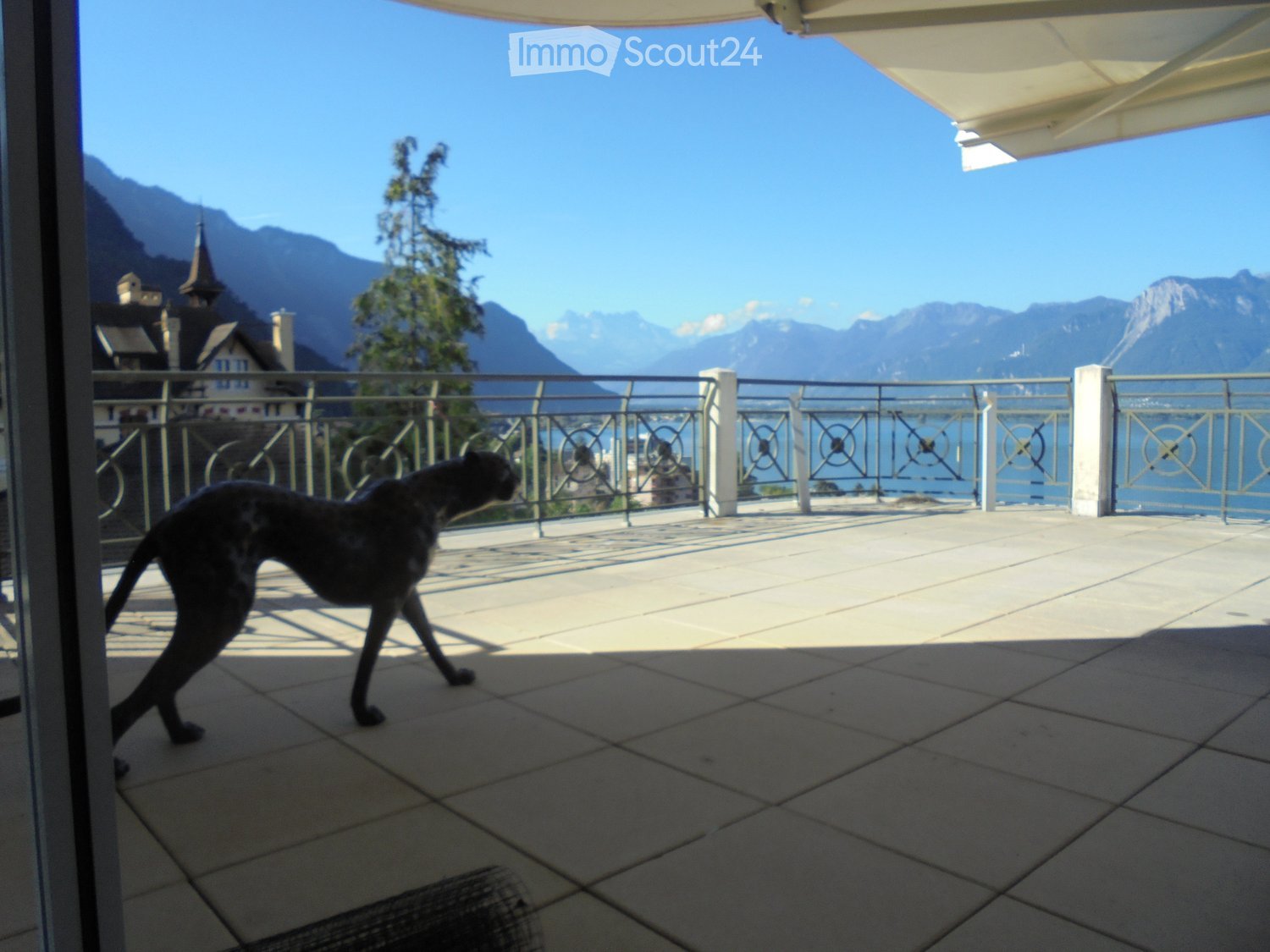 Balcony with a metal railing, overlooking a scenic lake and mountain landscape. A large cat is walking on the tiled floor.