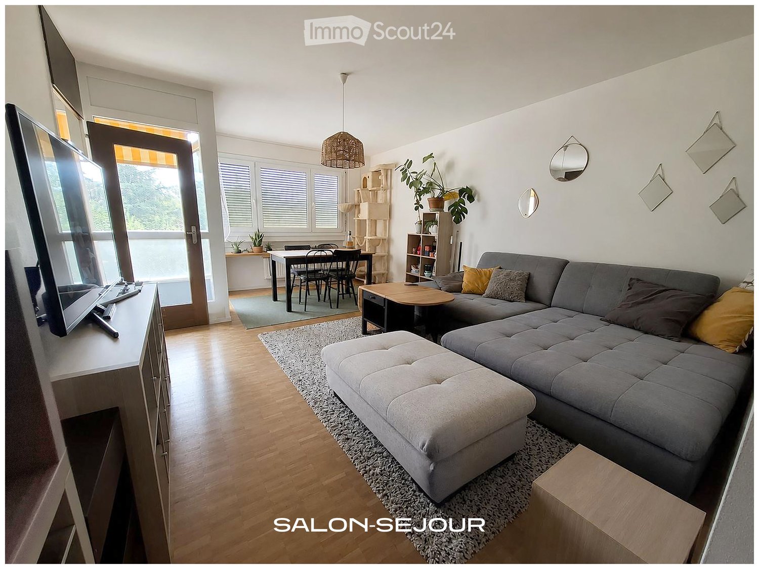 Modern living room with grey sectional sofa, ottoman, coffee table, TV stand, dining area with table and chairs, pendant light, and decorative mirrors on the wall.