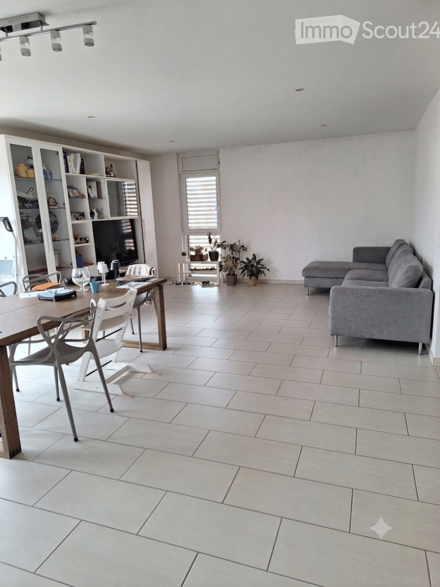 Bright living and dining area with white tiled flooring, grey sectional sofa, wooden dining table with chairs, bookshelf, small side table with potted plants, and modern lighting fixtures.