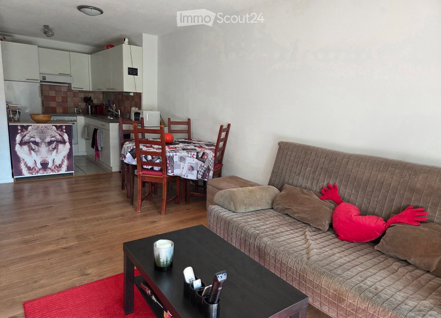 A modern living room with a sofa, coffee table, dining area, and kitchen. The room features wooden flooring, a red rug, and a decorative pillow on the sofa.