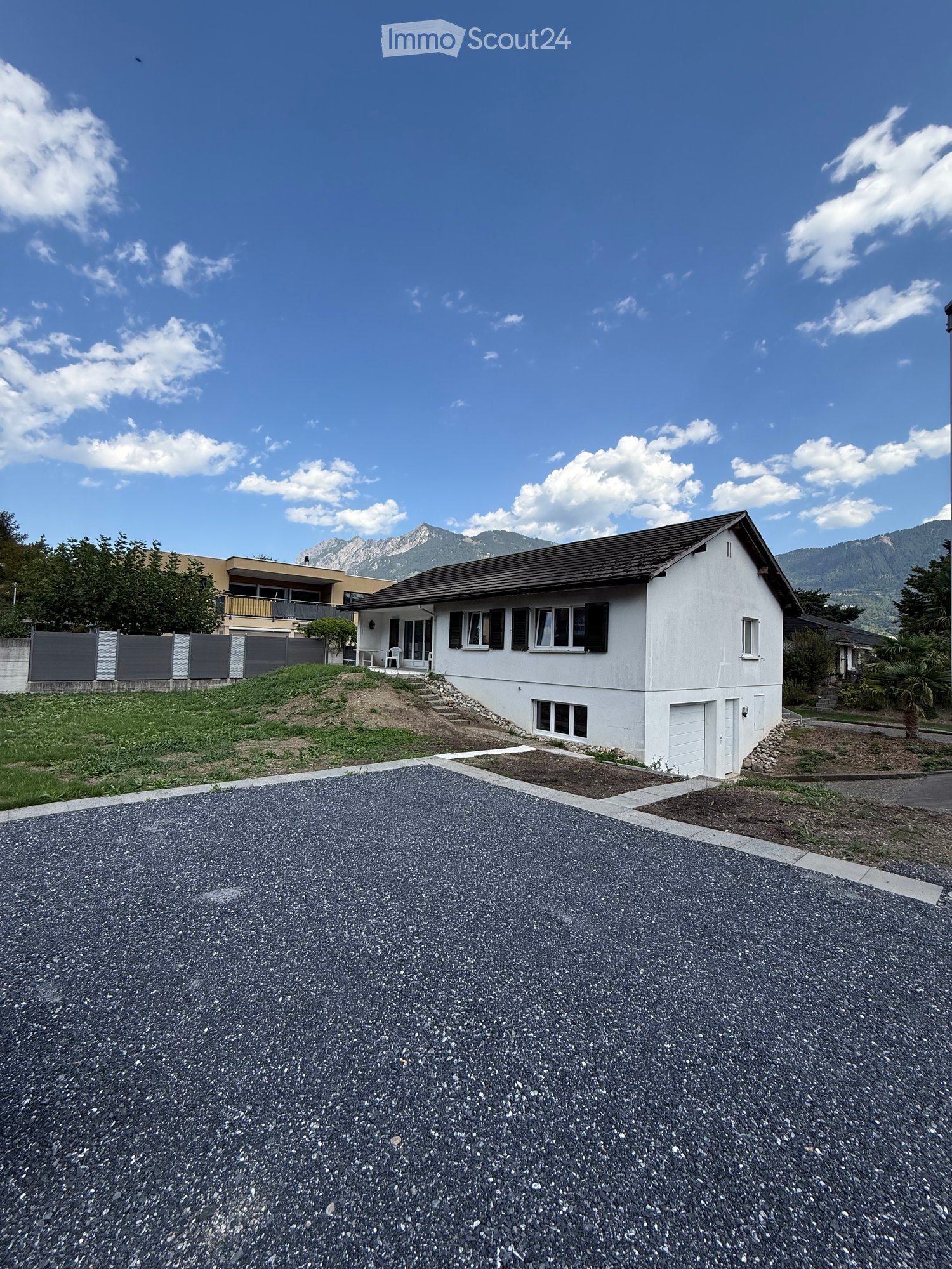 Single-story white house with a dark roof, multiple windows, and a garage. The house is situated on a raised ground floor with a gravel driveway and a small garden area.