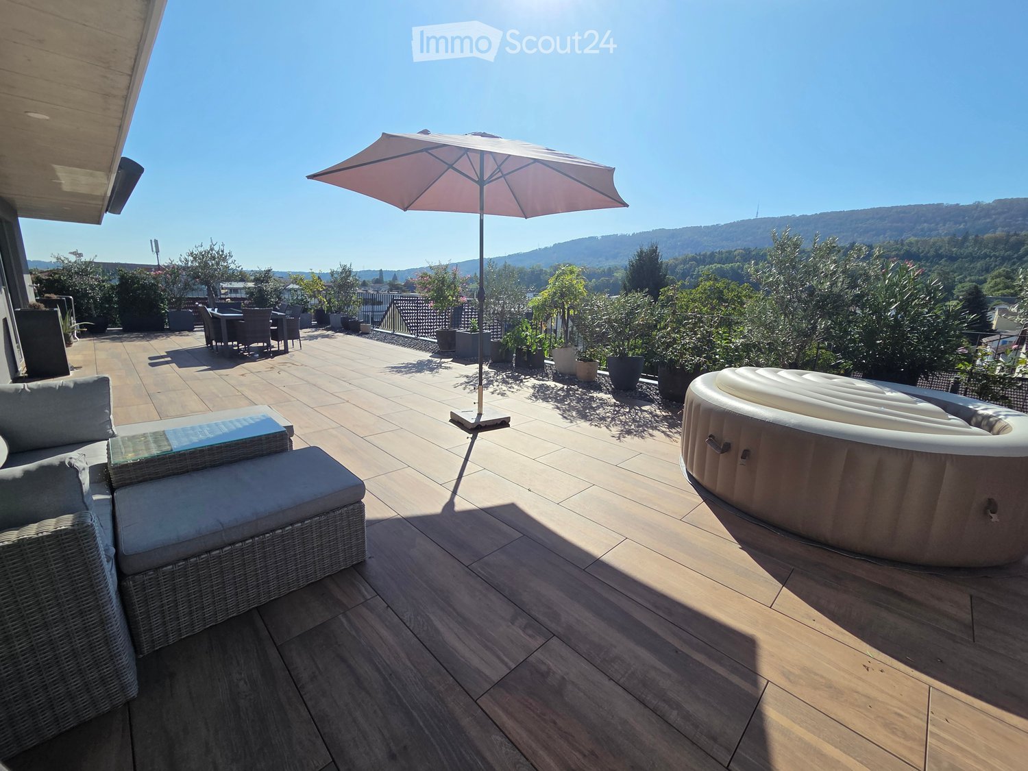 A spacious wooden deck with a sofa, table, chairs, umbrella, and a hot tub, overlooking a scenic valley.