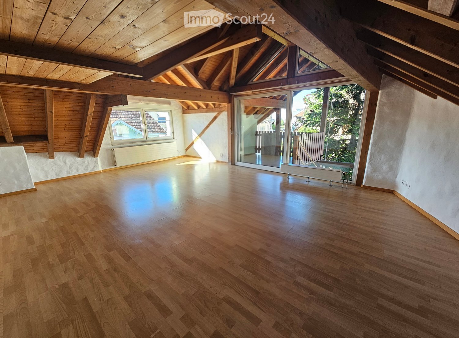 Empty attic room with wooden floors, exposed wooden beams, large windows, and a sliding glass door leading to a balcony.