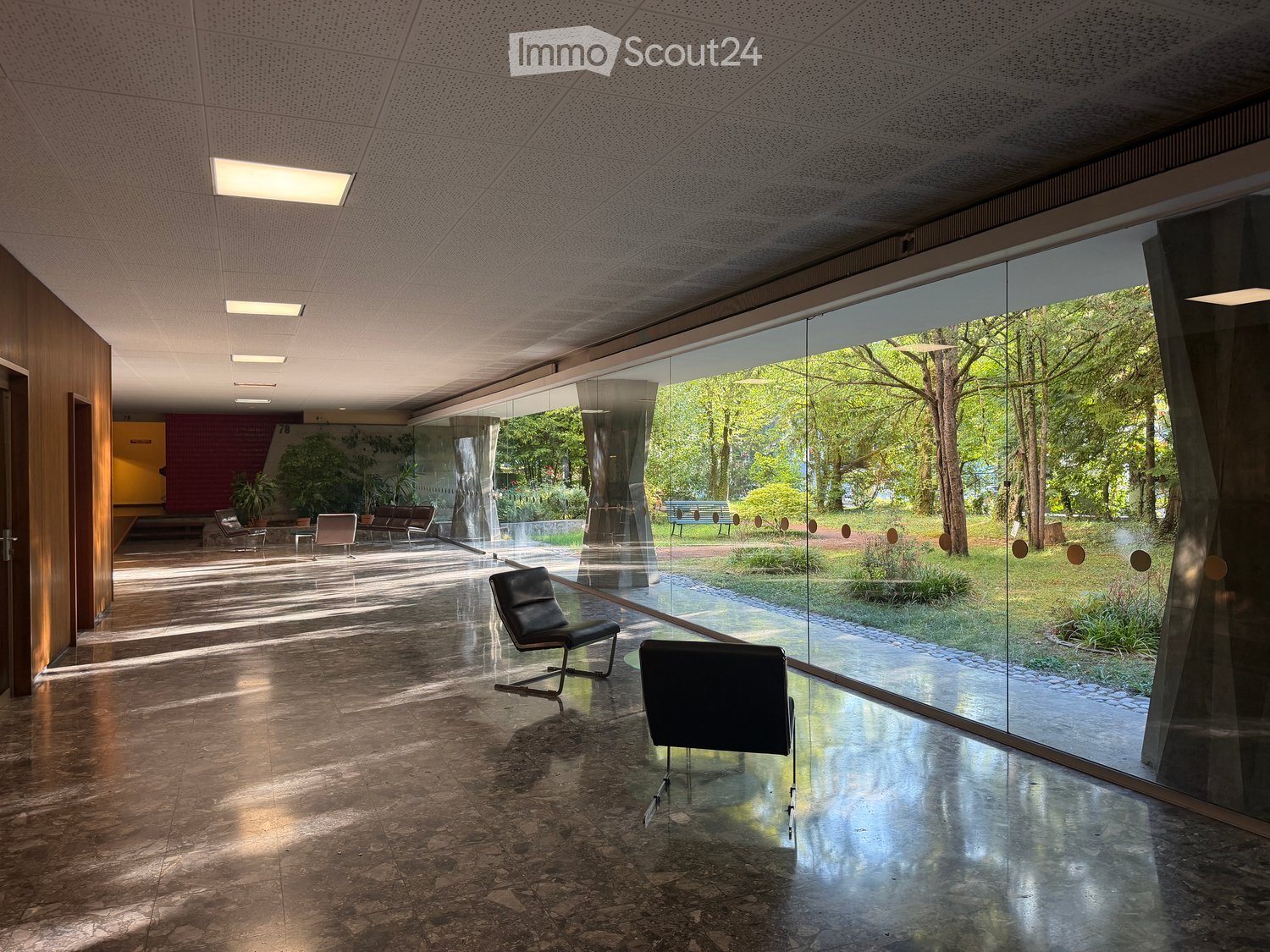 Modern, spacious room with large glass walls offering a view of a lush garden. Features include sleek black chairs, a polished stone floor, and recessed lighting.