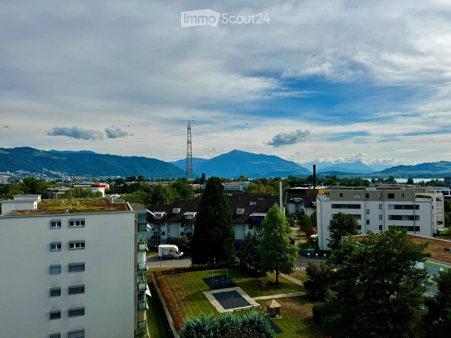 A view of a residential area with multiple apartment buildings, green spaces, and a playground, with mountains in the background.