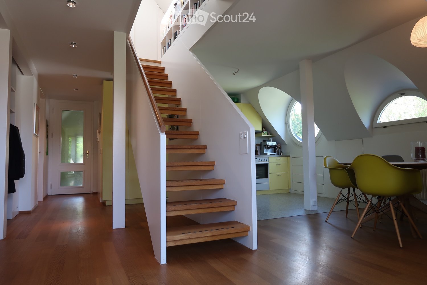 Modern hallway with wooden flooring, staircase leading upwards, built-in shelving, and a small kitchen area visible to the right.