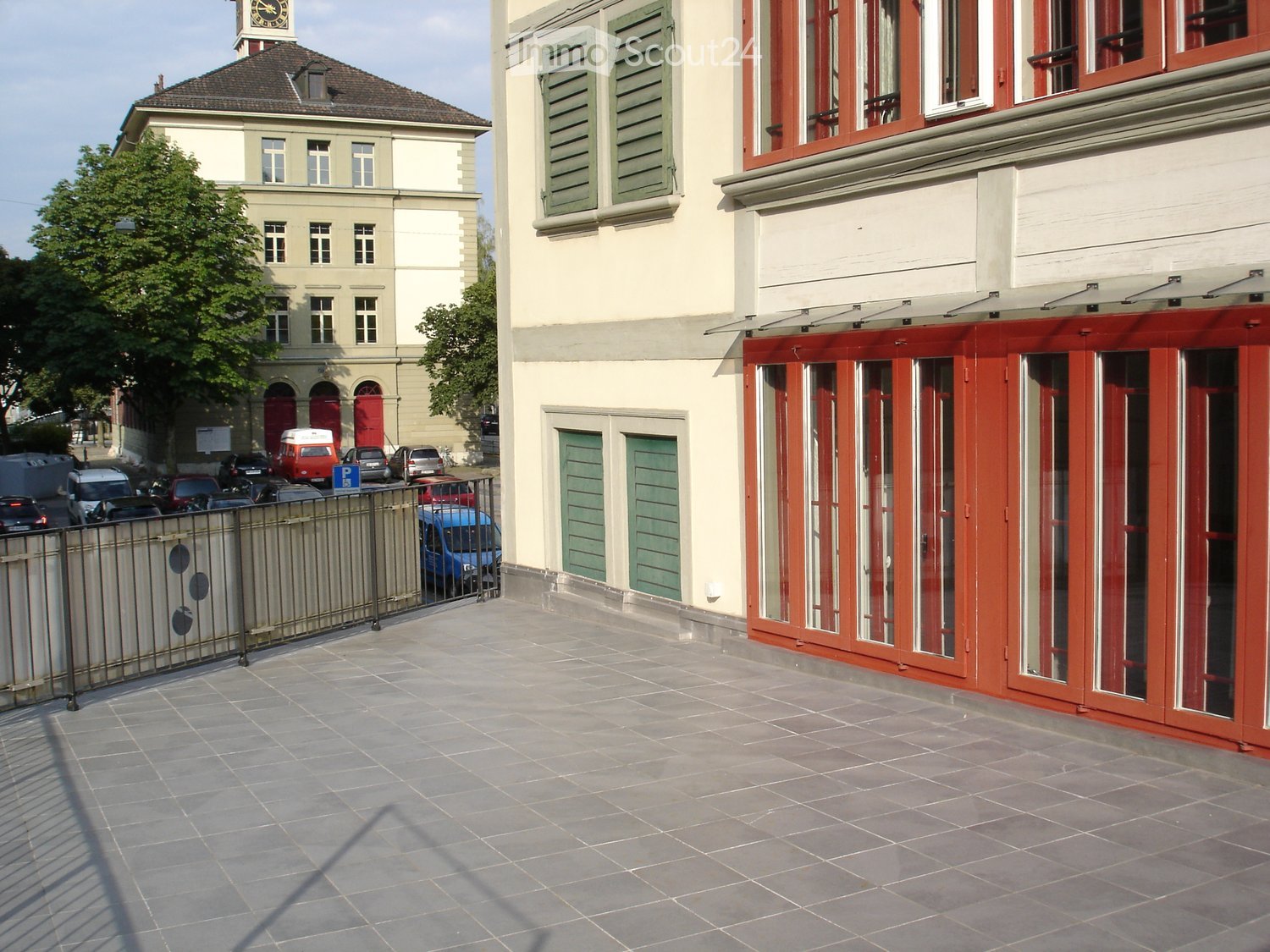 A multi-story building with a tiled roof, green shutters, and a clock tower in the background. The foreground shows a paved area with a metal fence and several parked cars.
