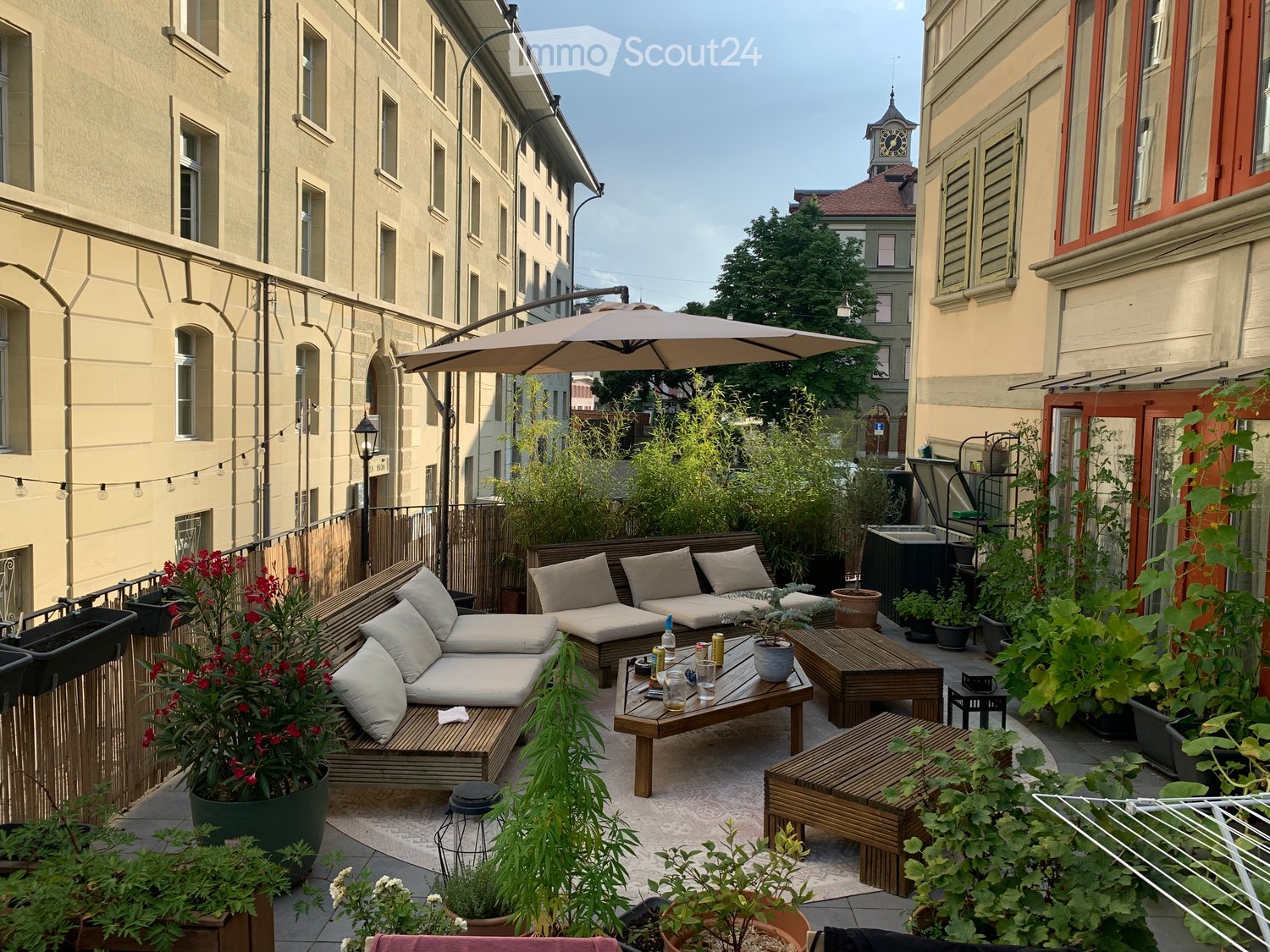 Cozy rooftop terrace with wooden seating, cushions, small table, potted plants, and an umbrella, overlooking a cityscape with historic buildings.
