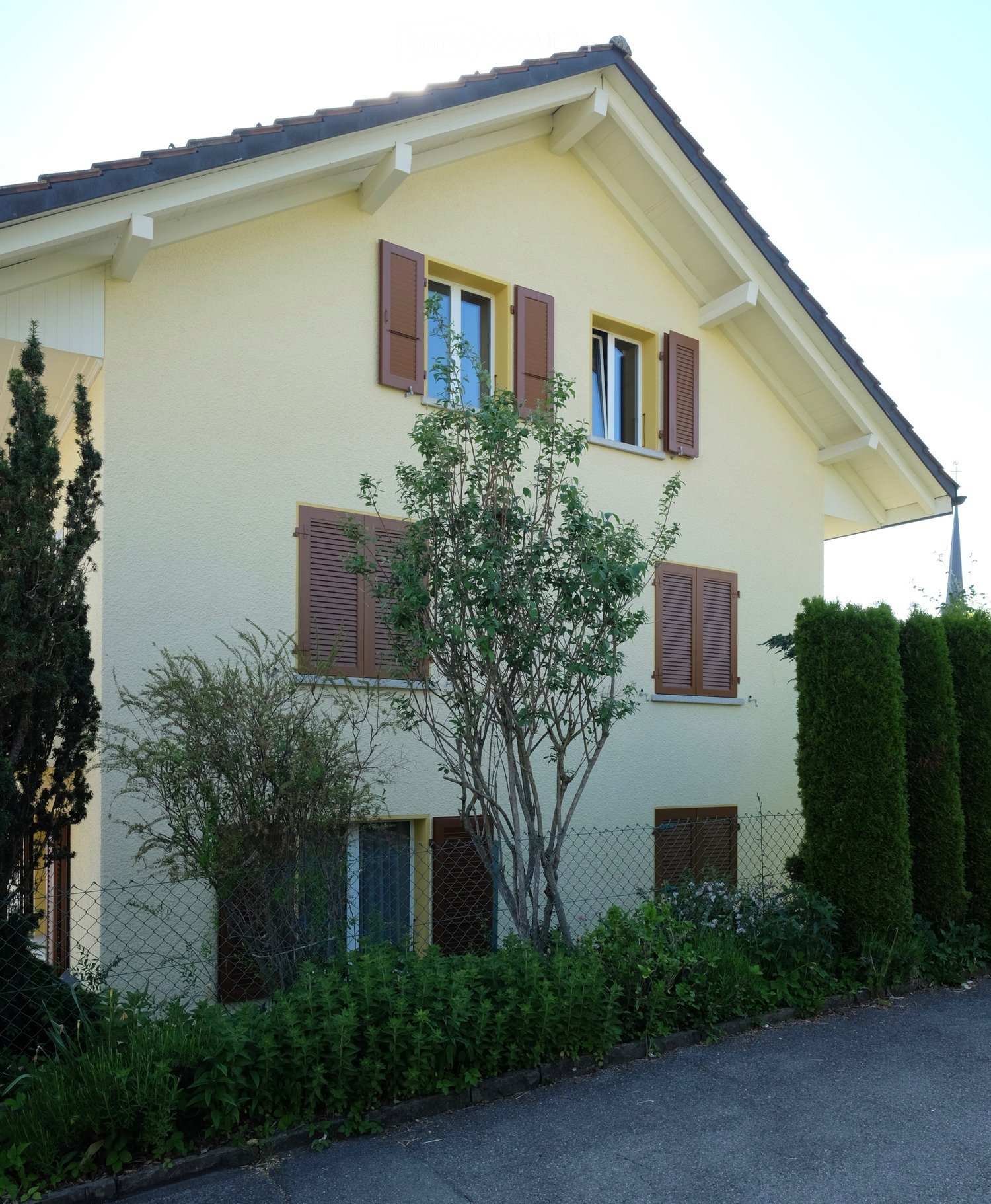 Two-story house with light yellow walls, dark brown shutters, and a pitched roof. The house is surrounded by a low fence, small trees, and shrubs.