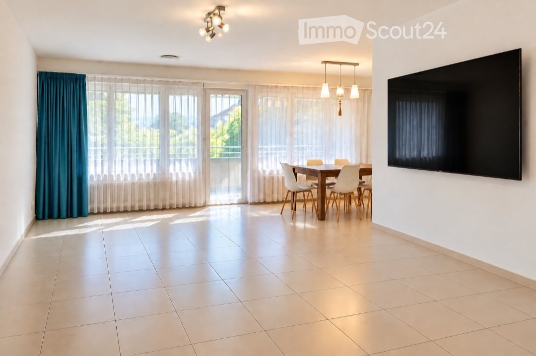 Bright dining area with a wooden dining table, white chairs, large windows with sheer curtains, and a flat-screen TV mounted on the wall.