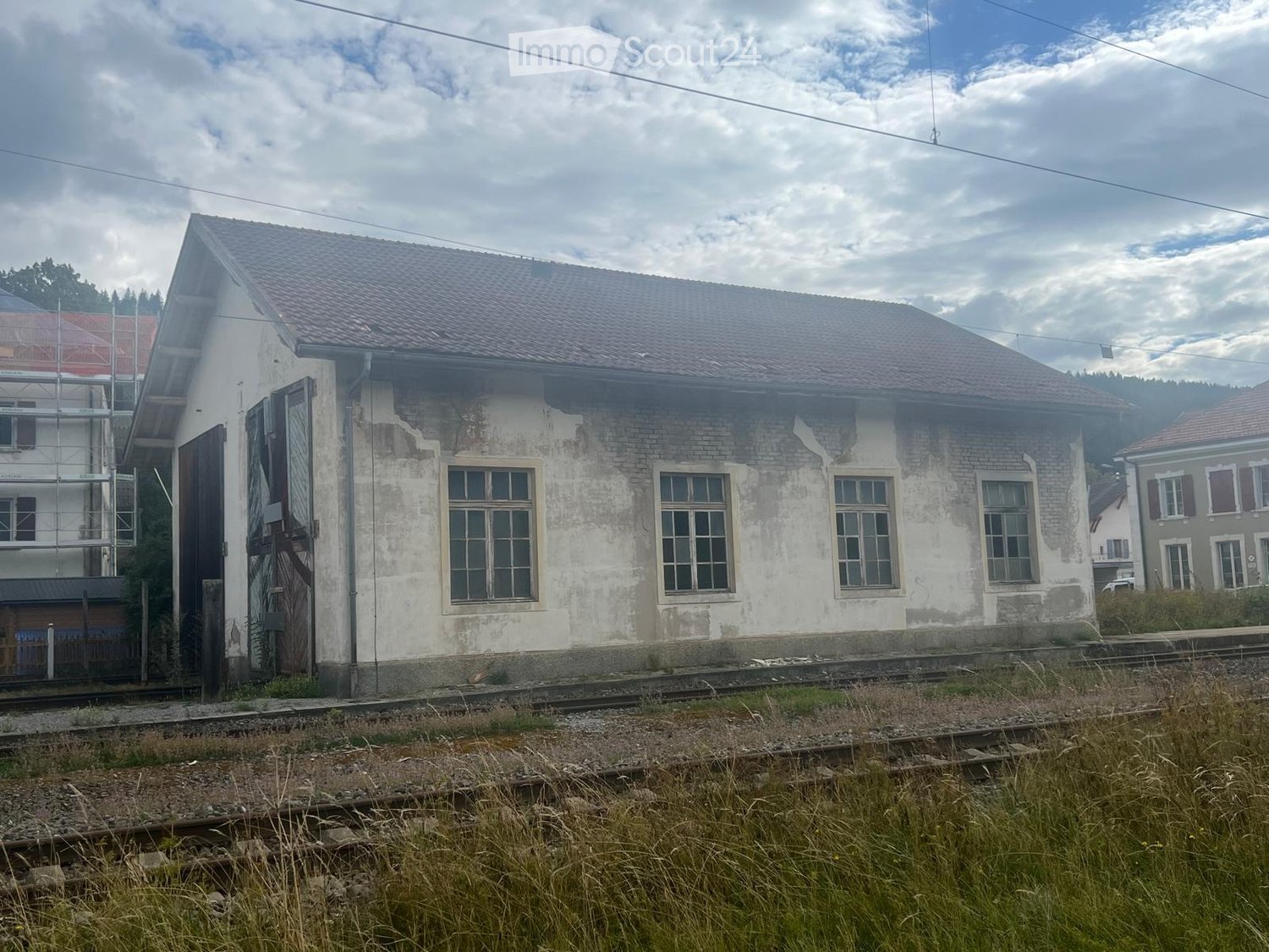 Two-story building with a tiled roof, brick facade, and multiple windows. The building appears to be in a rural setting with railway tracks in the foreground.