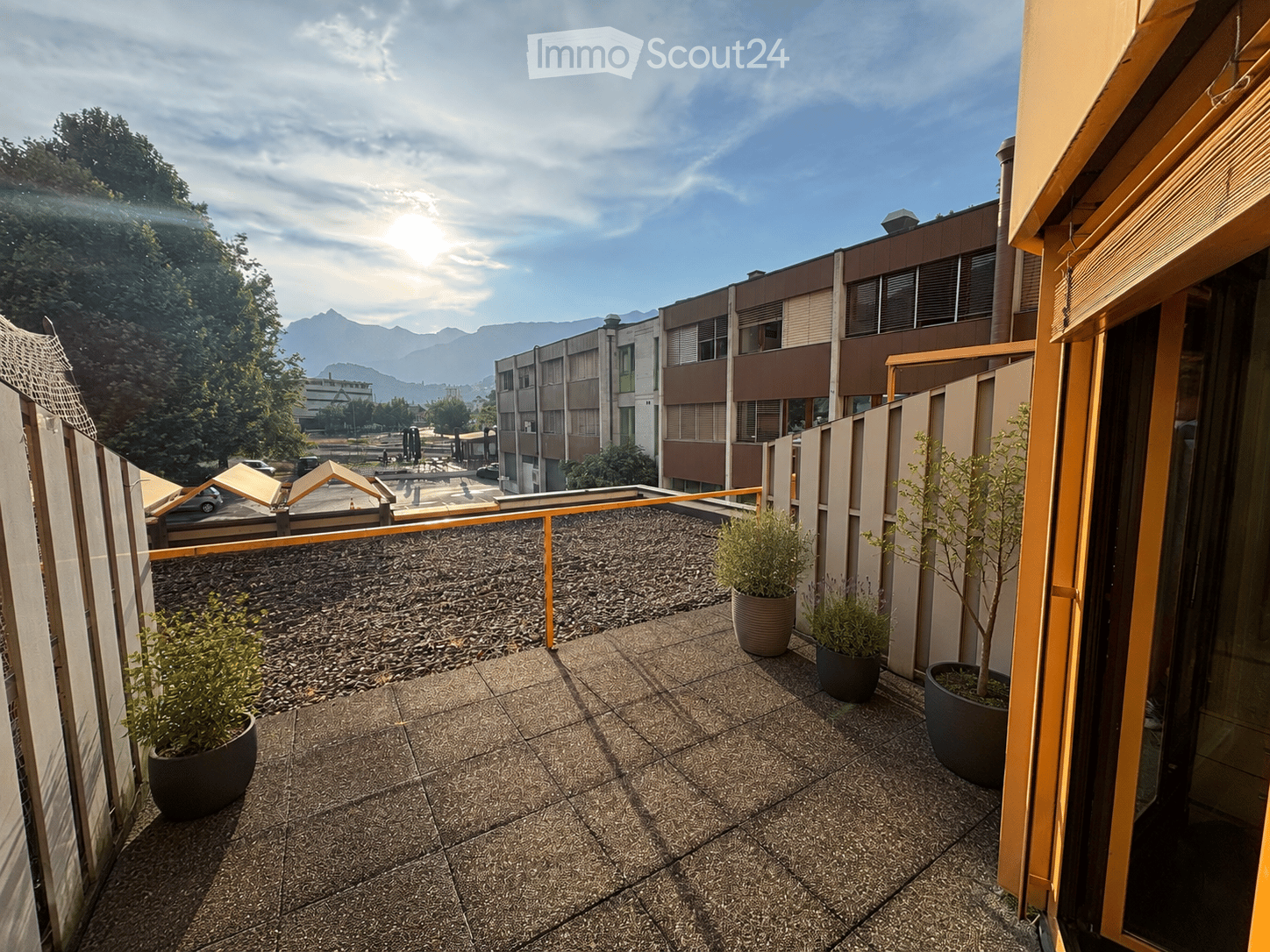 Balcony with tiled floor, potted plants, and a view of surrounding buildings and mountains.