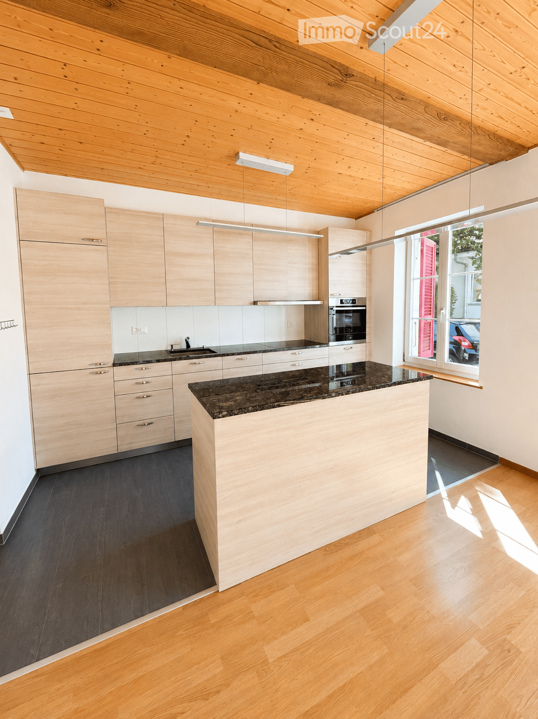 Modern kitchen with light wooden cabinets, dark granite countertop, and a central island. Features include a sink, oven, and pendant lighting.