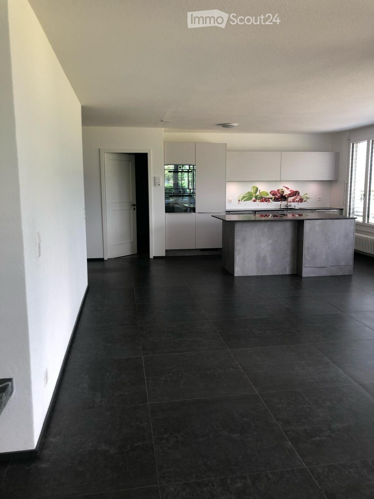 Modern kitchen with dark tiled flooring, white cabinets, and a central island. The space is well-lit with natural light from large windows and has a sleek, contemporary design.