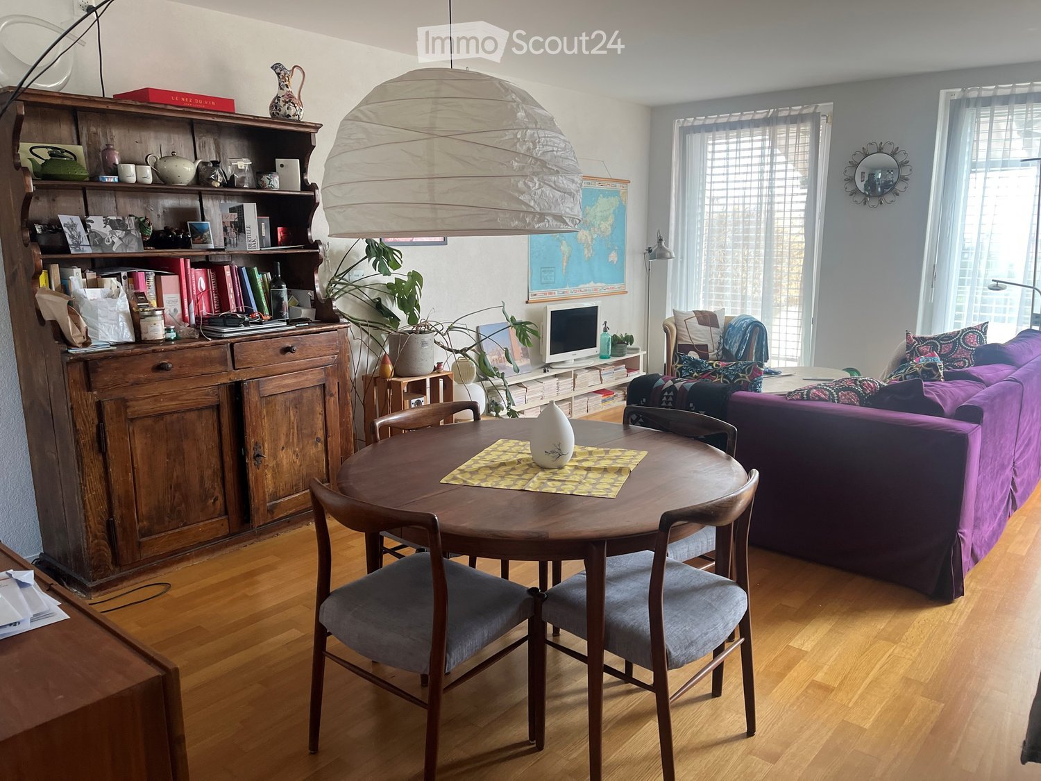 A cozy living room with a purple sofa, wooden dining table with chairs, a wooden cabinet, a hanging lamp, and a large window with blinds.