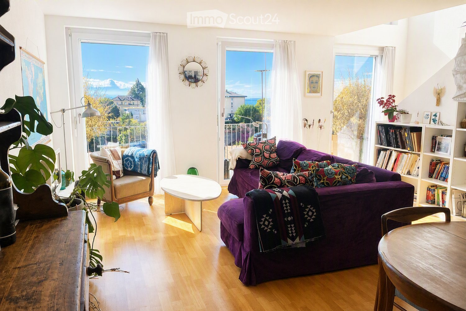 Bright living room with wooden flooring, large windows offering a scenic view, a purple sofa with colorful pillows, a wooden coffee table, a bookshelf, and a small seating area with a chair and plant.