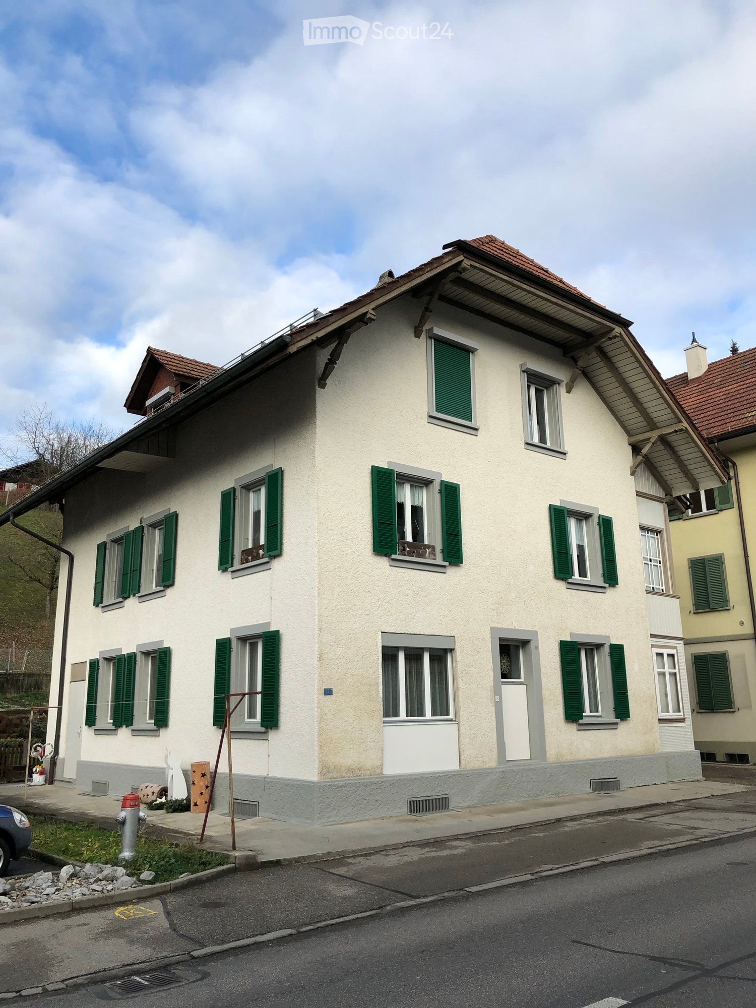 Two-story house with light beige walls, green shutters, multiple windows, and a red-tiled roof.