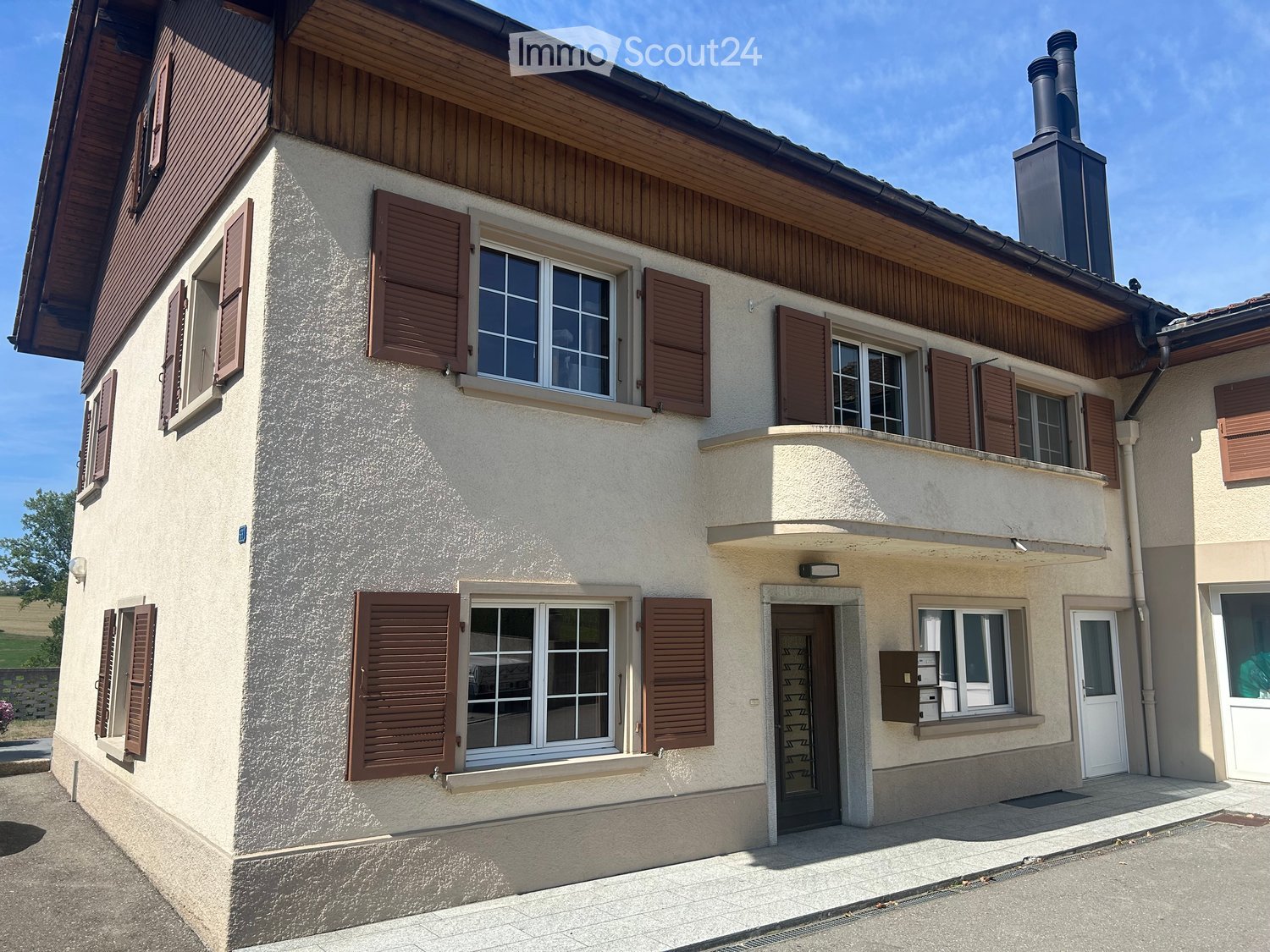 Two-story house with beige walls, wooden shutters, and multiple windows. Features a small balcony on the upper floor and a chimney on the roof.