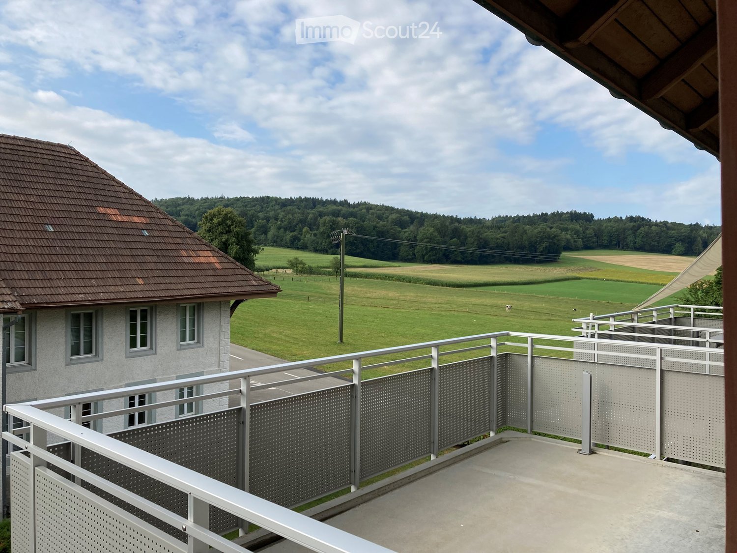 Balcony with metal railing overlooking a green field and a house with a brown roof.