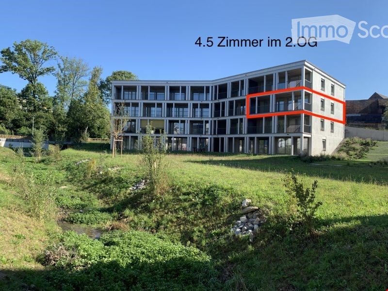Modern multi-story apartment building with balconies, surrounded by green space and a small stream.