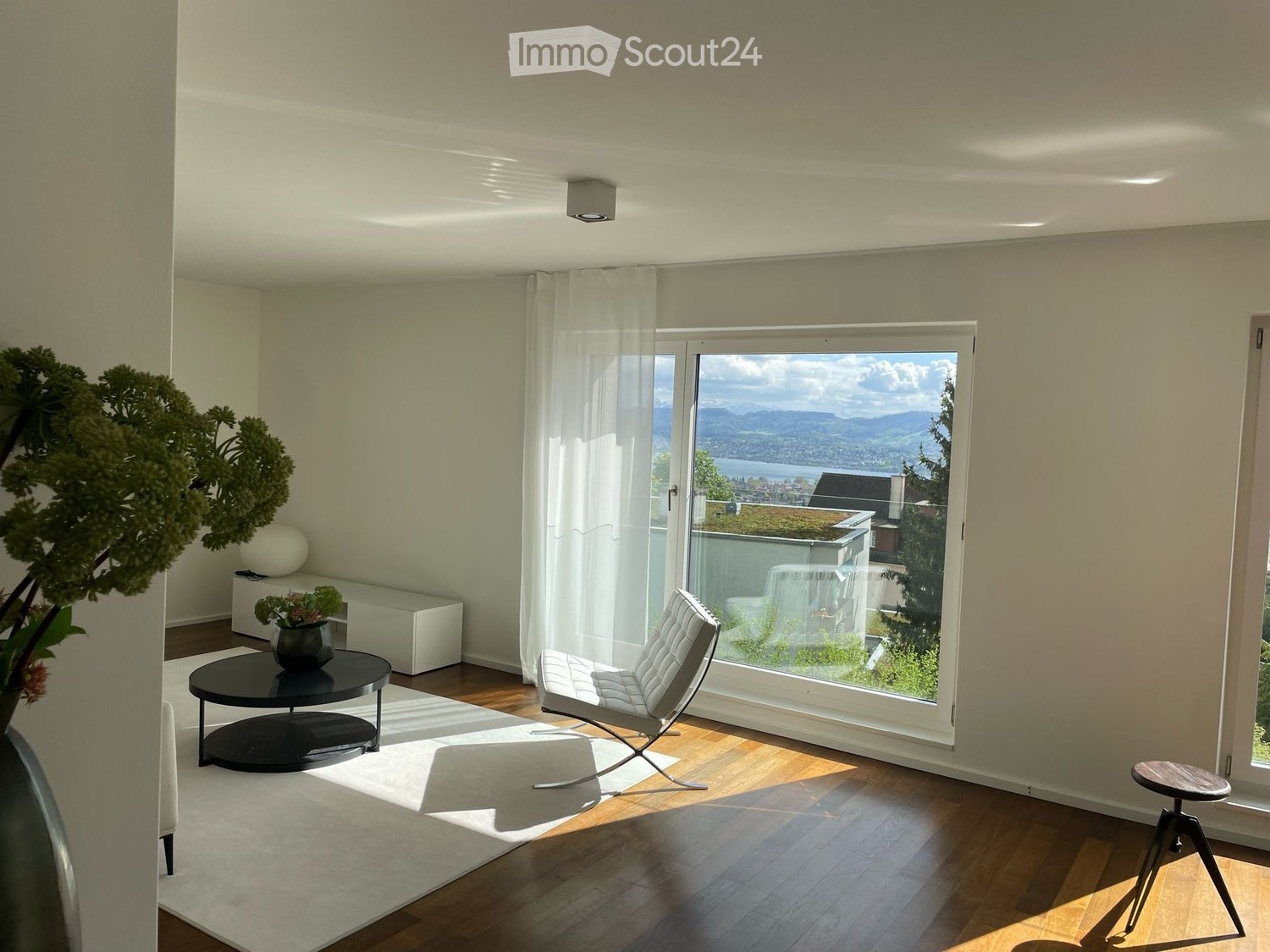 Modern living room with large windows offering a scenic view, featuring a white leather sofa, a black coffee table, a small stool, and decorative vases.