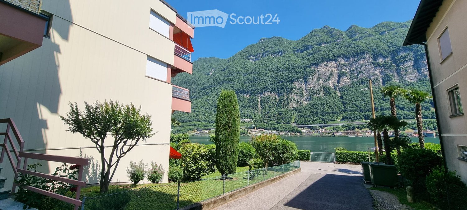 A multi-story residential building with balconies overlooking a scenic lake and mountain backdrop. The area includes greenery, trees, and a paved pathway.