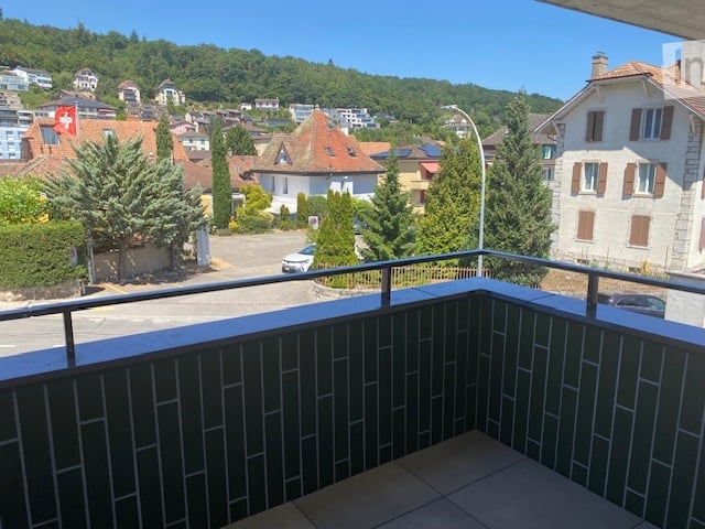 Balcony with a view of a town, featuring tiled flooring and a metal railing, overlooking a street with parked cars and surrounding buildings.