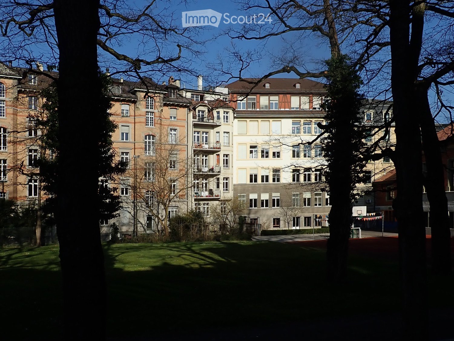 A multi-story residential building with multiple windows and balconies, surrounded by trees and a grassy area.