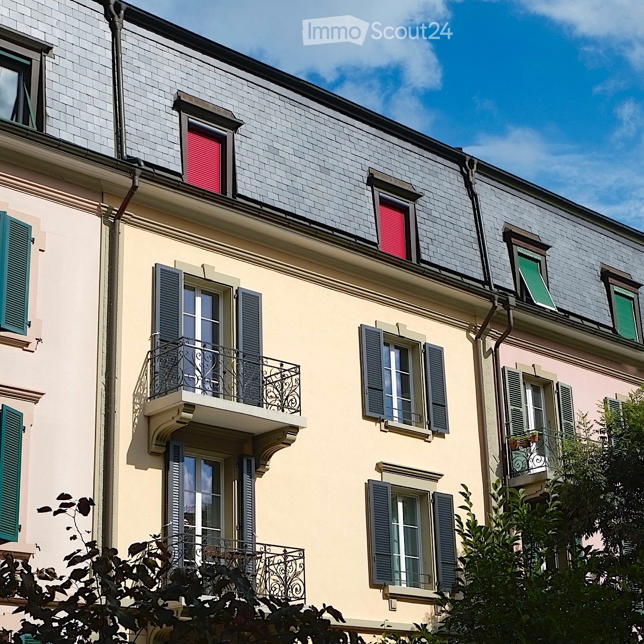 A multi-story residential building with light yellow walls, multiple windows with shutters, and ornate balconies with metal railings.