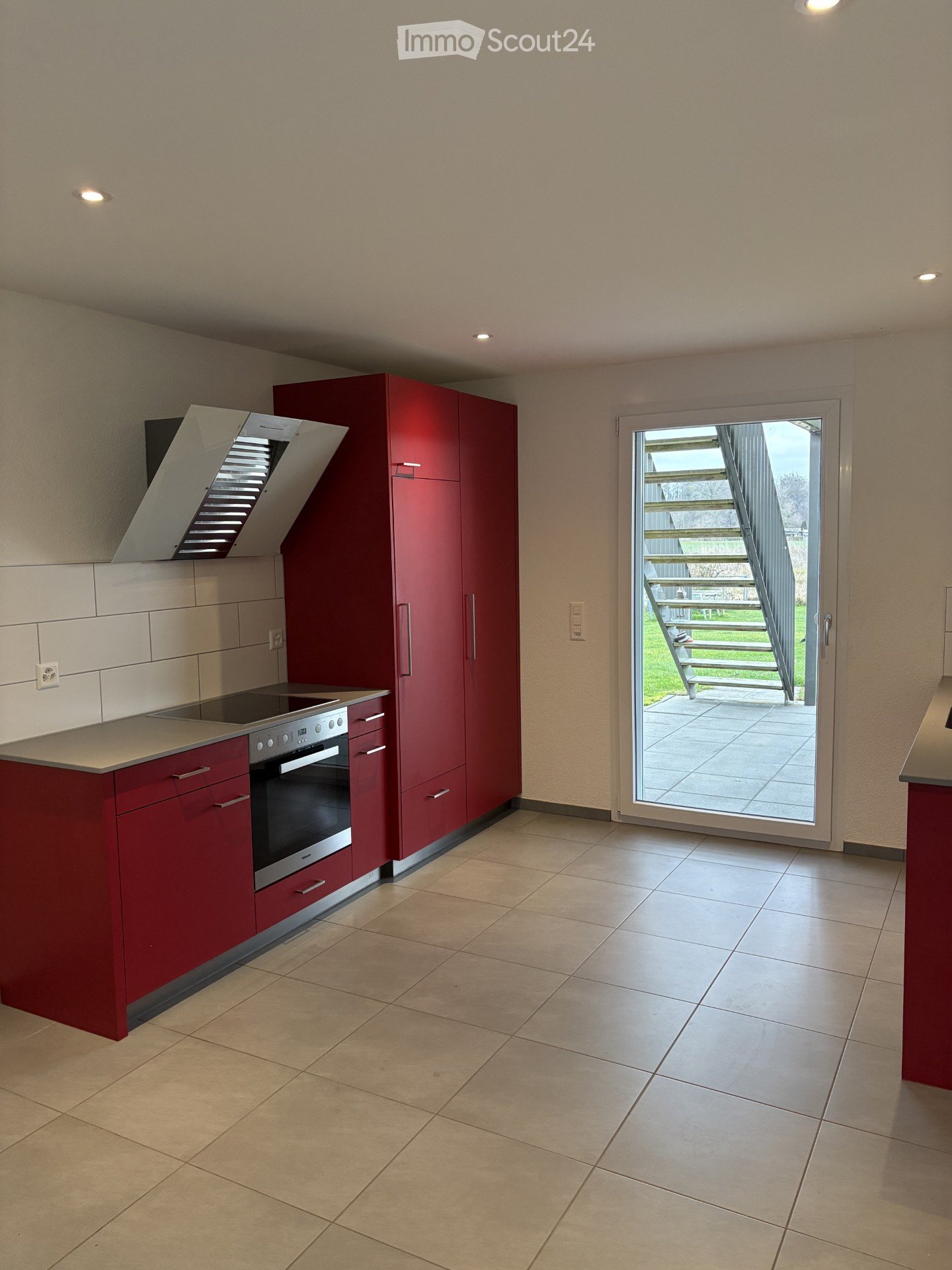 Modern kitchen with red cabinets, stainless steel oven, range hood, and a sliding glass door leading to an outdoor area.