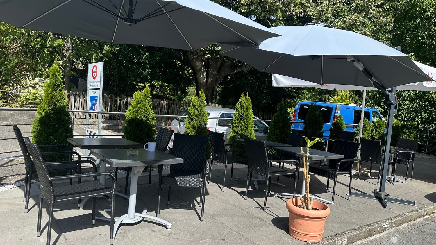 Outdoor seating area with tables, chairs, umbrellas, and a potted plant, surrounded by greenery and parked vehicles.