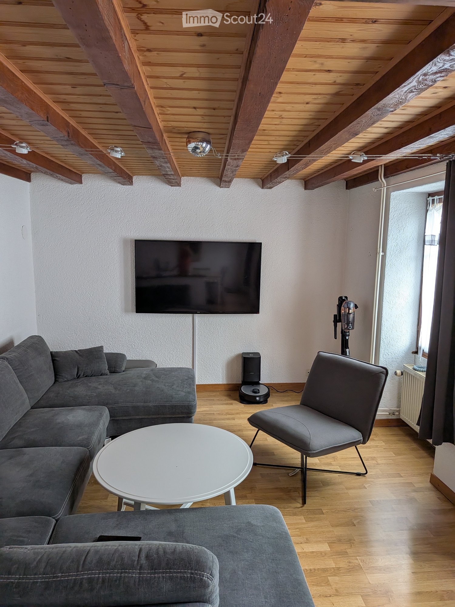 Modern living room with exposed wooden beams, grey sofa, white coffee table, grey armchair, flat-screen TV, and a robotic vacuum cleaner.