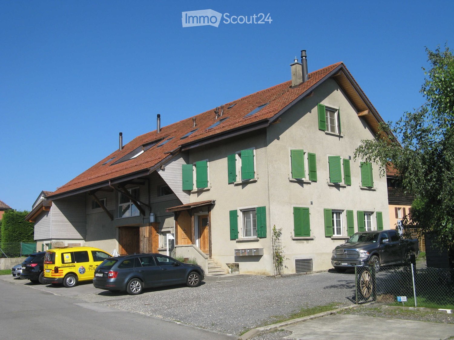 Two-story house with beige walls, green shutters, red-tiled roof, and multiple chimneys. The house has a covered entrance and is surrounded by a gravel driveway with several parked cars.