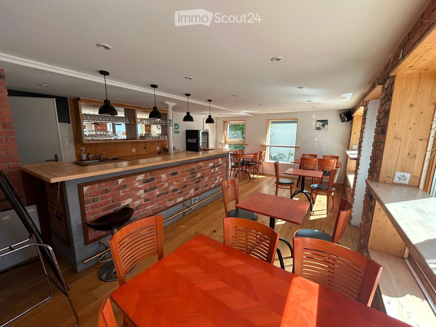 A cozy bar area with wooden tables, chairs, a bar counter, and a refrigerator, illuminated by hanging lights.