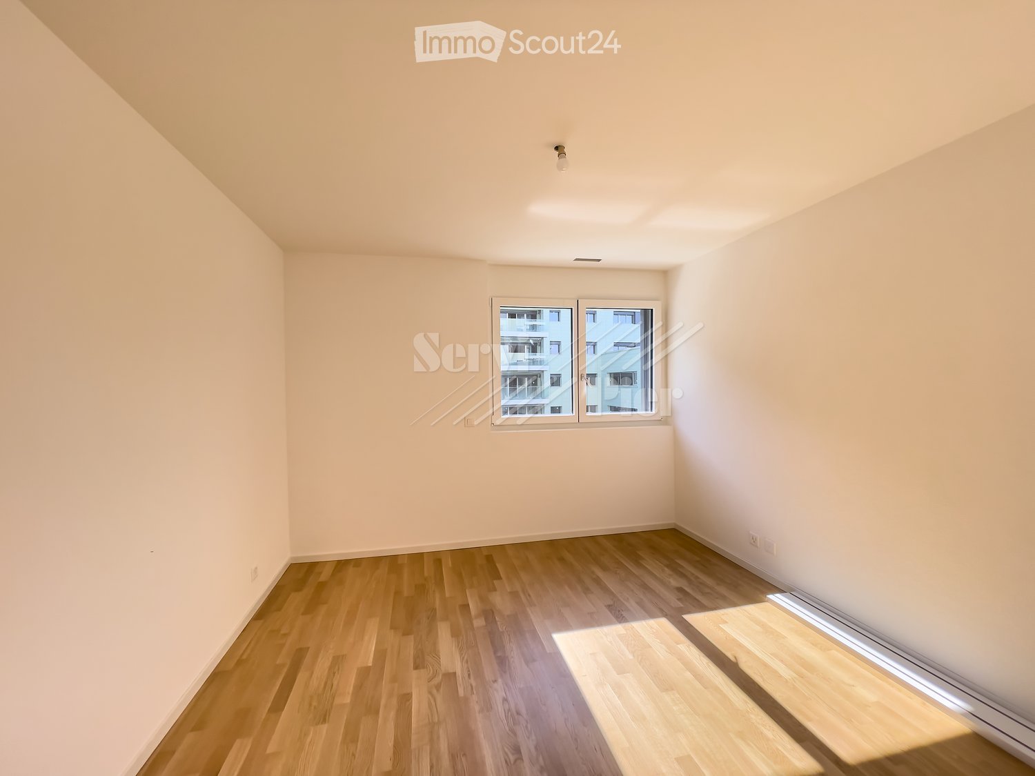 Empty room with wooden flooring, white walls, and a large window offering a view of neighboring buildings.