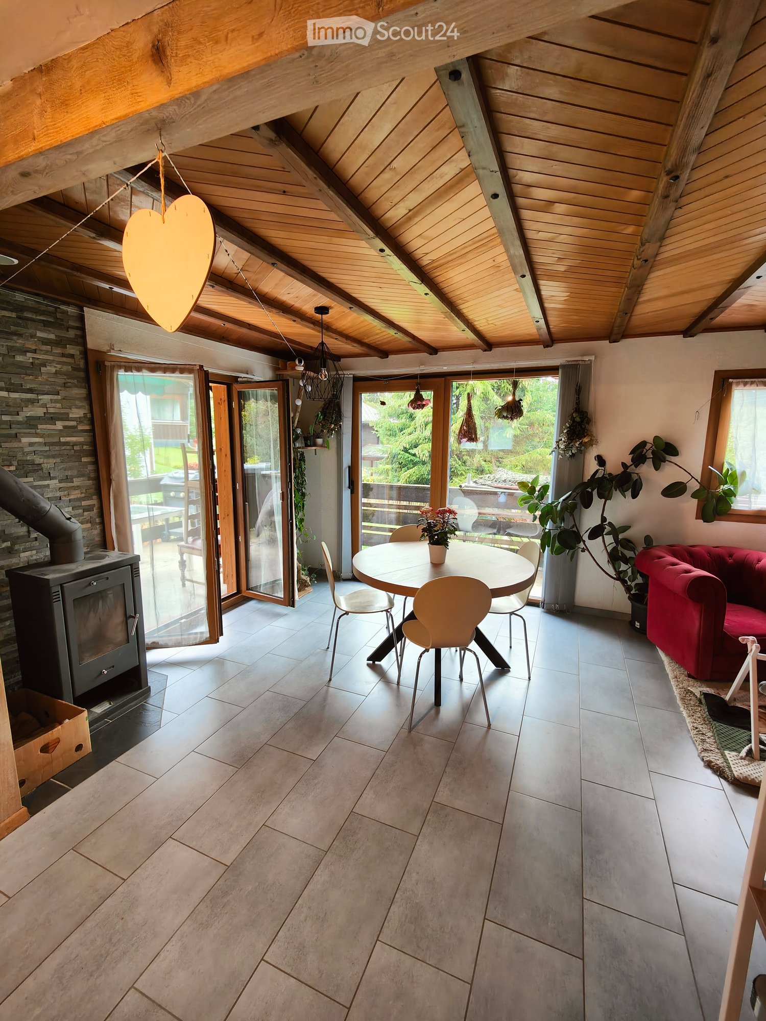 Bright living space with exposed wooden beams, a wood stove, a red sofa, a dining table with chairs, and large windows opening to a garden.