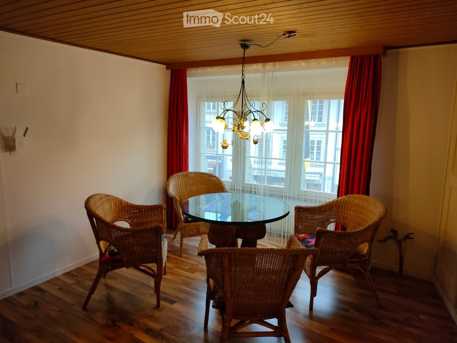 Dining area with a glass table and four wicker chairs, wooden flooring, large windows with red curtains, and a hanging chandelier.