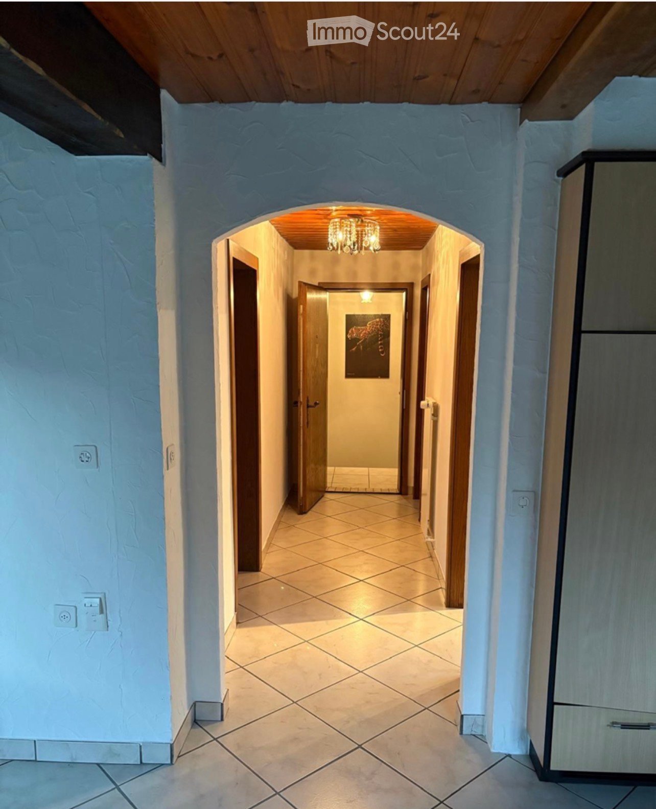 A well-lit hallway with tiled flooring, light blue textured walls, and wooden doors leading to other rooms. A chandelier hangs from the ceiling at the far end.