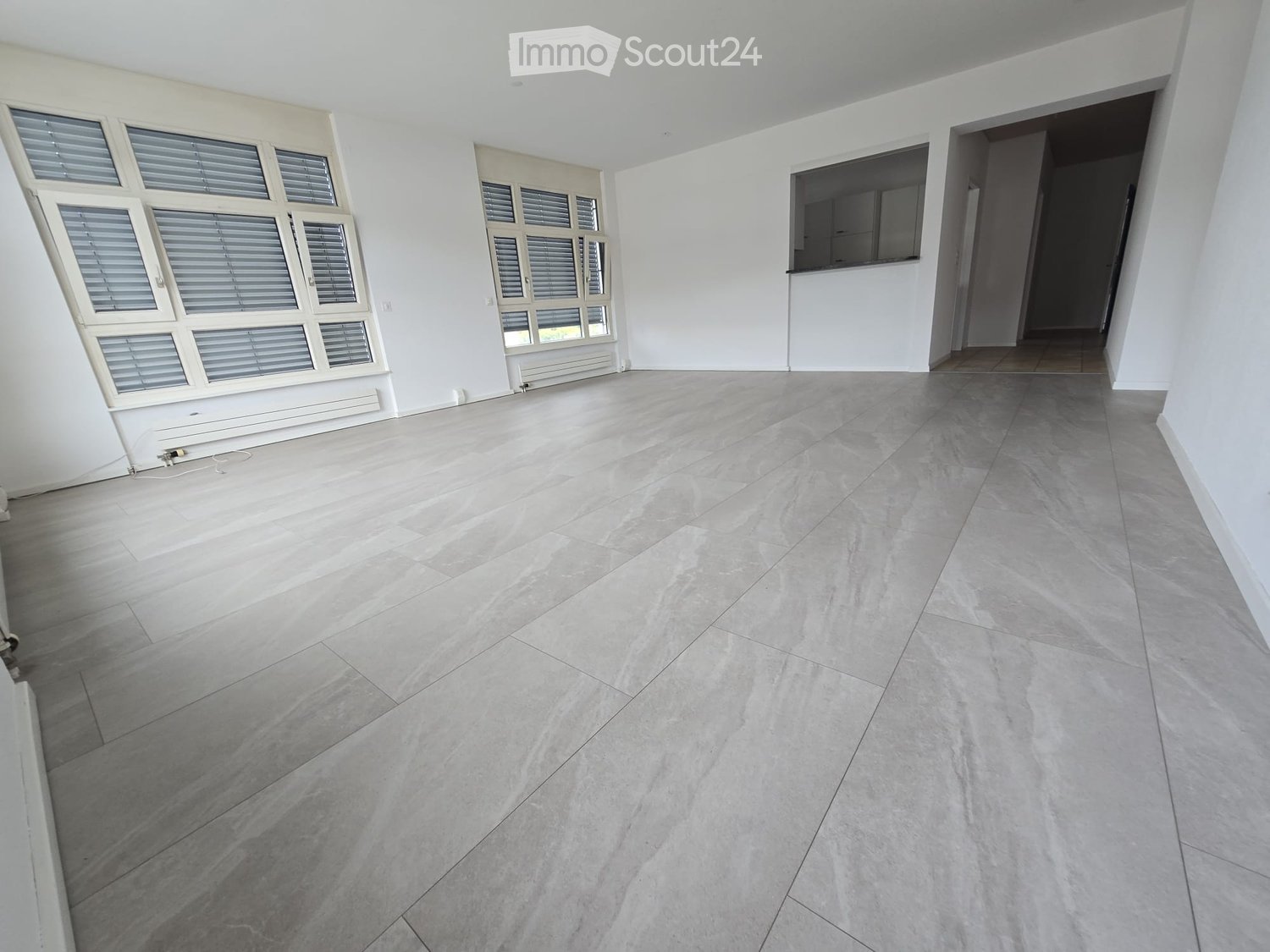 Empty room with light grey tiled floor, white walls, large windows with blinds, and a doorway leading to another space.