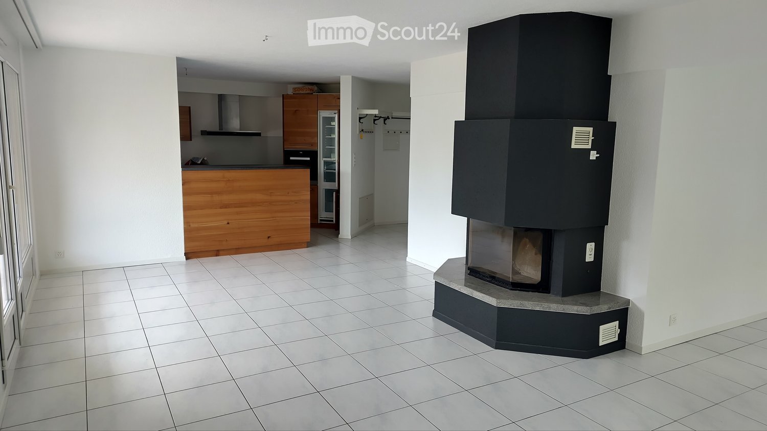 Modern living space with a built-in fireplace, white tiled floor, and an open kitchen area with wooden cabinets and a refrigerator.
