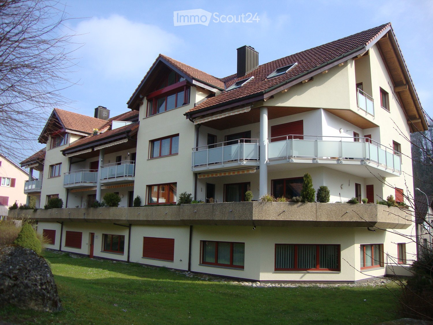 Two-story house with beige exterior, red shutters, multiple balconies, and a well-maintained lawn.