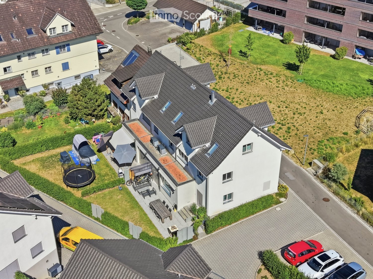 Aerial view of a residential area with multiple houses, gardens, and parked cars. Features include a trampoline, outdoor furniture, and a well-maintained lawn.