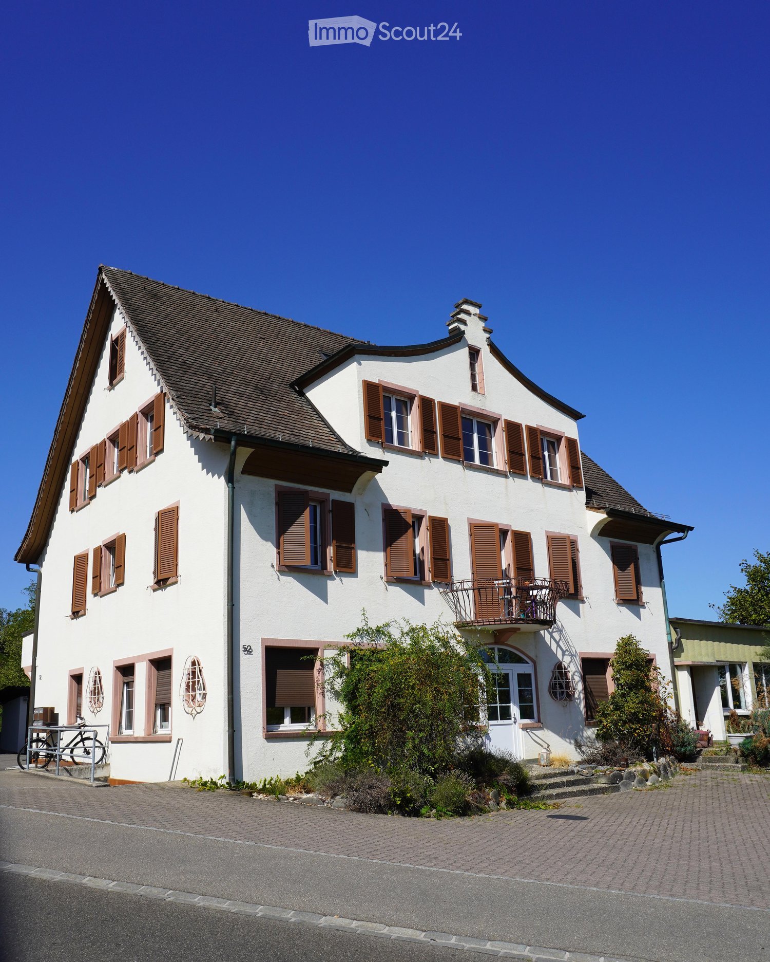 Two-story house with white walls, brown shutters, and a tiled roof. Features include a balcony on the upper floor, a small garden area, and a paved driveway.
