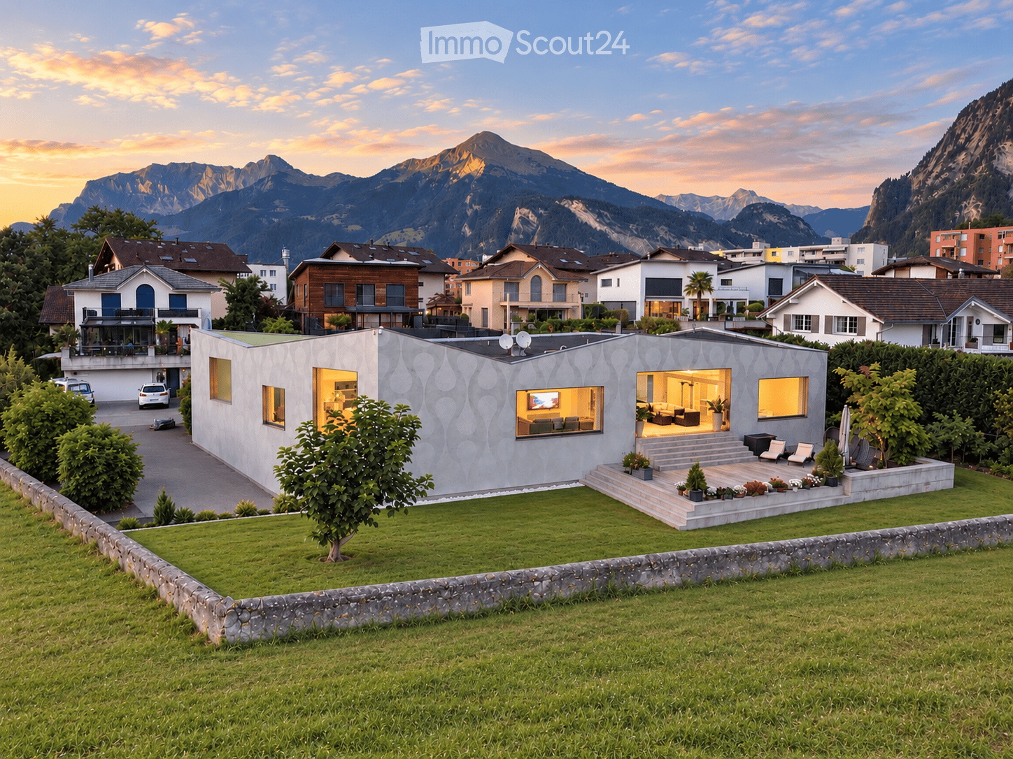 Modern single-story house with a textured facade, large windows, and a well-maintained lawn in a scenic mountain setting.