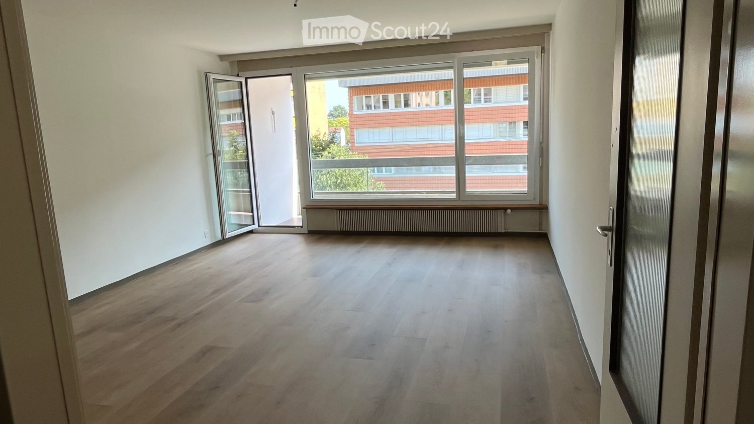 Empty room with wooden flooring, white walls, large window, and open sliding door leading to a balcony.