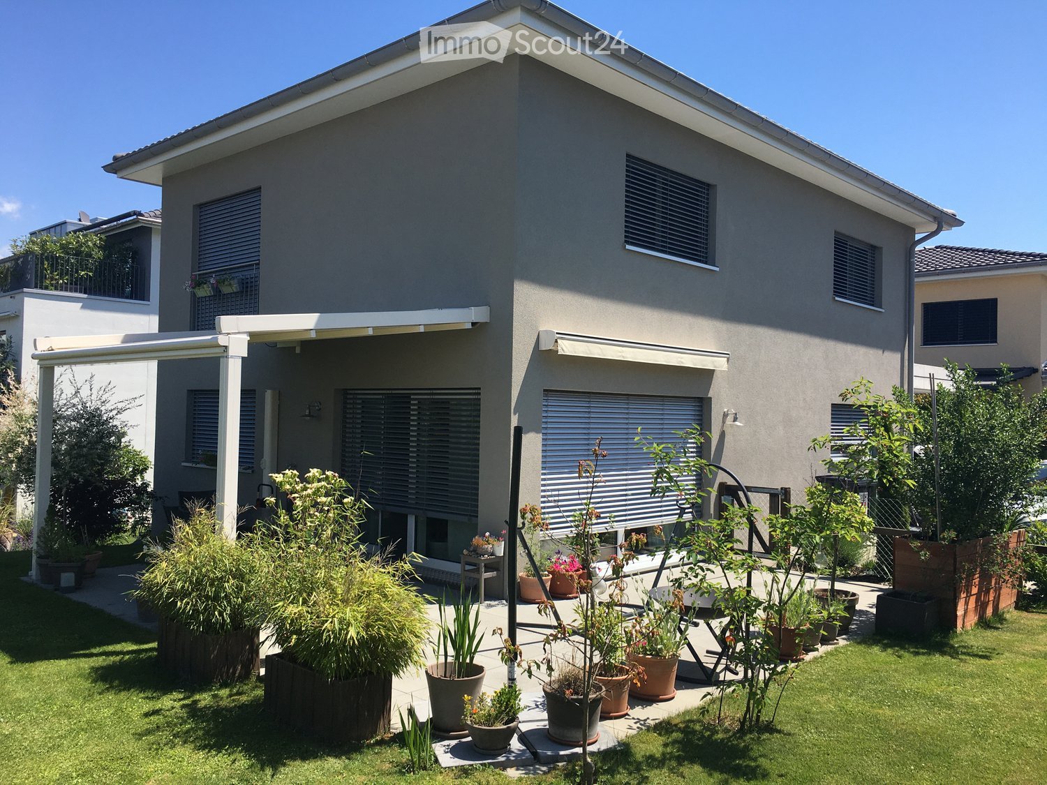 Modern two-story house with grey exterior, large windows, covered patio area, and well-maintained garden with potted plants and lawn.