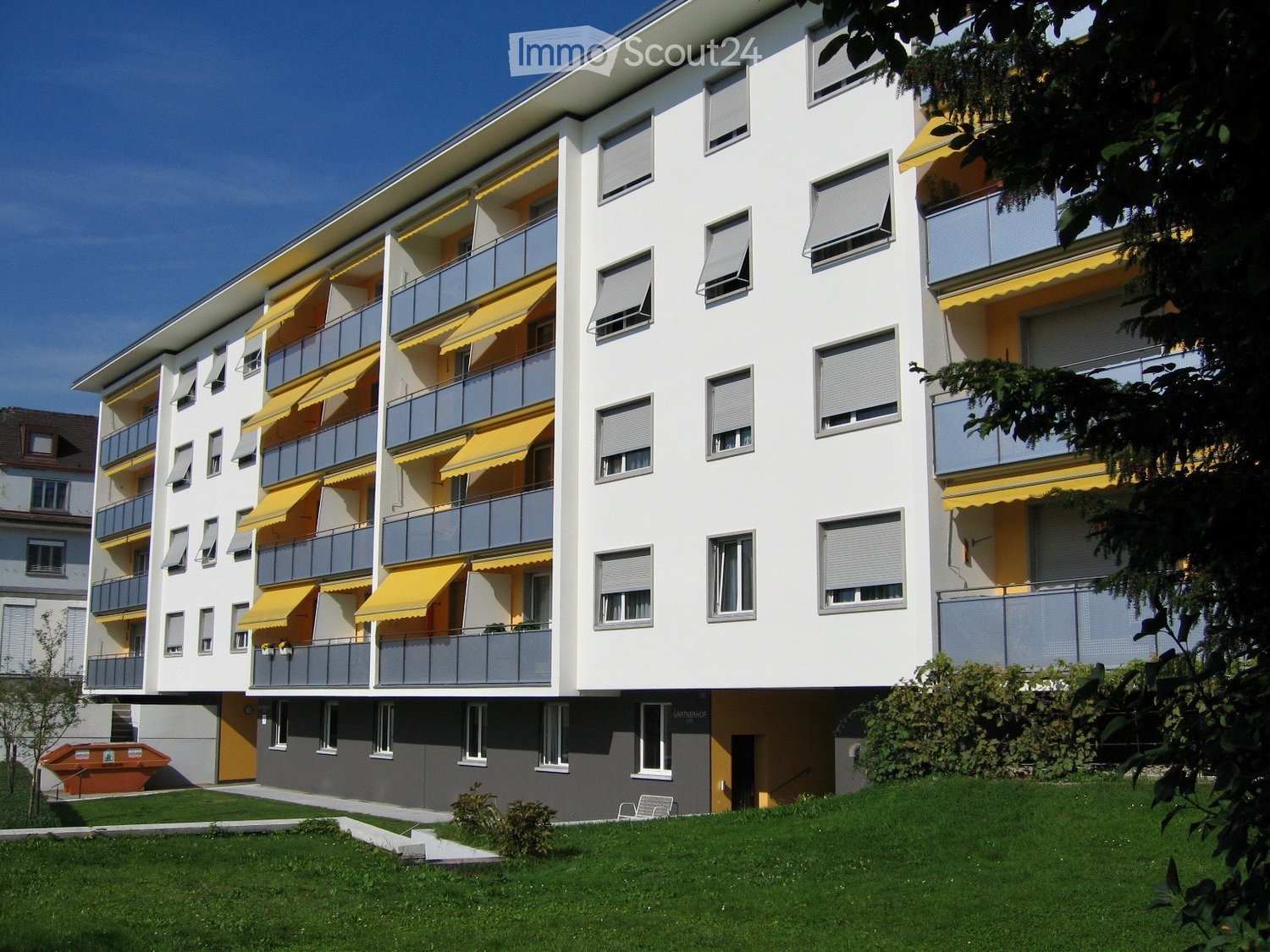 Modern multi-story apartment building with white and yellow facade, multiple balconies with yellow awnings, and a well-maintained lawn in front.