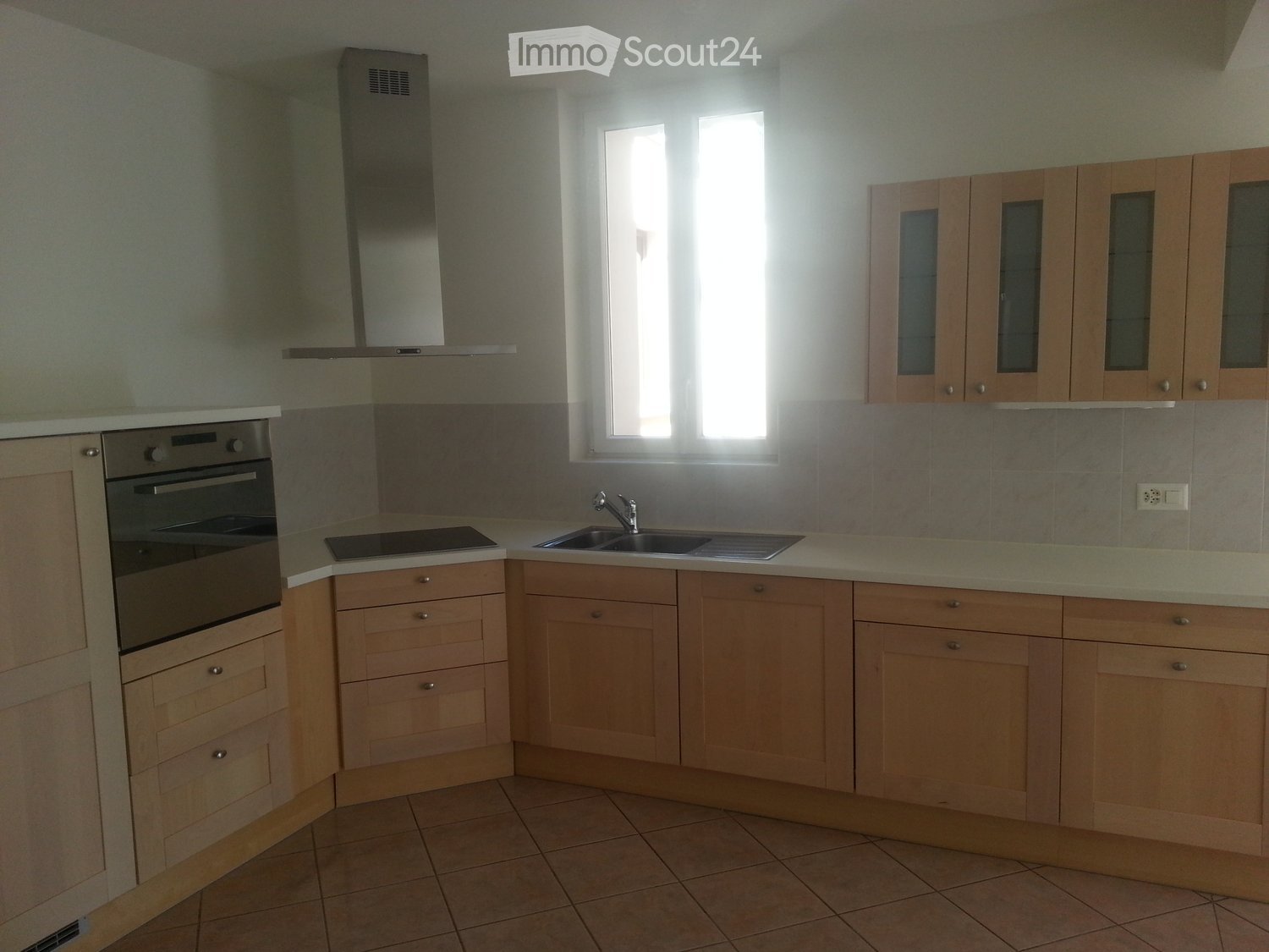 Modern kitchen with light wooden cabinets, stainless steel oven, sink, and range hood. Features a tiled backsplash and tiled floor.
